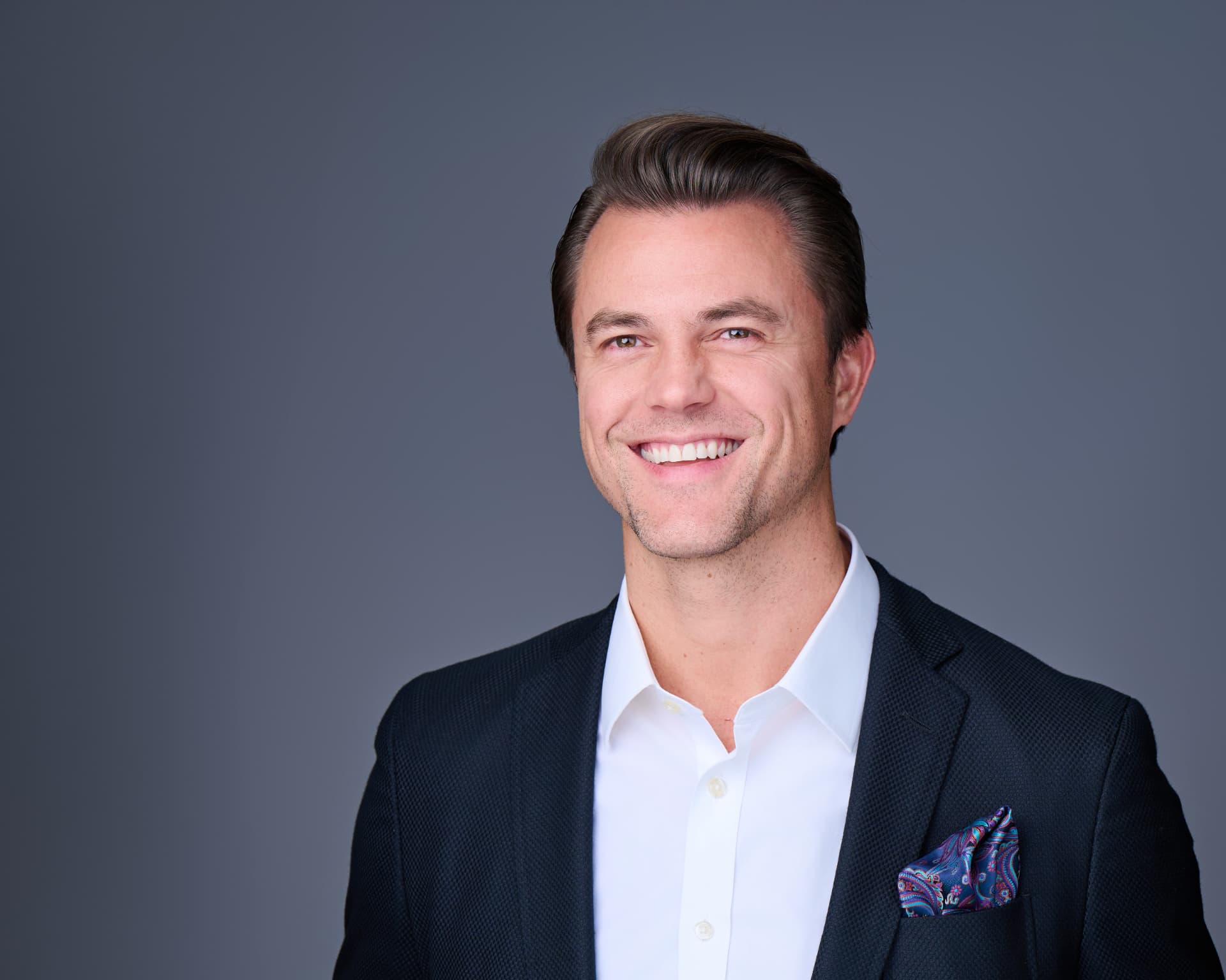 Smiling man in a navy blazer with a patterned pocket square against a gray studio backdrop