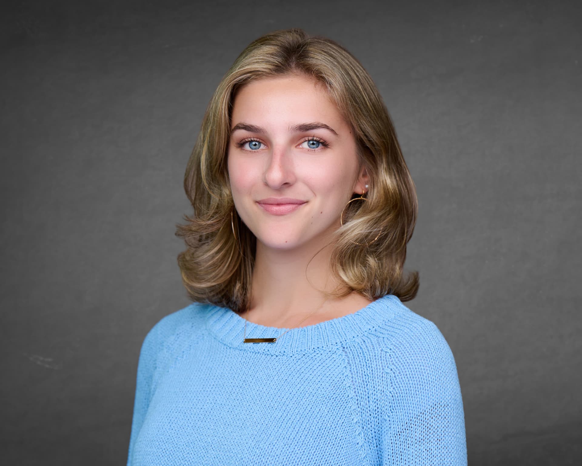 Professional headshot of a smiling woman in a sky blue sweater against a charcoal gray backdrop
